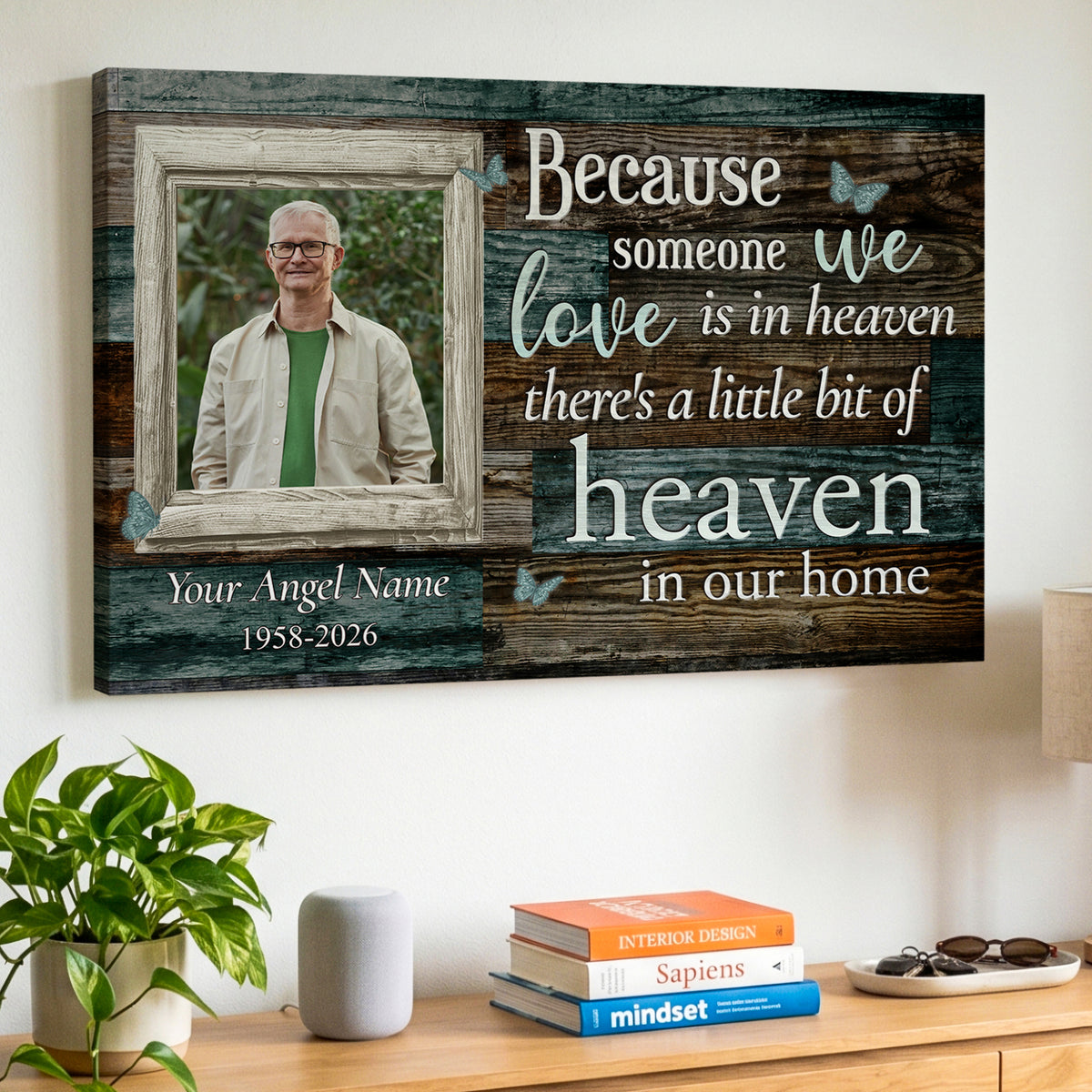 A personalized wooden wall canvas print hanging on a white wall above a wooden console table. The canvas features a rustic wood-grain background with white and light blue text that reads 'Because someone we love is in heaven there's a little bit of heaven in our home', along with a customizable photo frame containing a portrait of a deceased loved one.