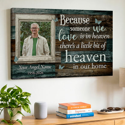 A personalized wooden wall canvas print hanging on a white wall above a wooden console table. The canvas features a rustic wood-grain background with white and light blue text that reads 'Because someone we love is in heaven there's a little bit of heaven in our home', along with a customizable photo frame containing a portrait of a deceased loved one.
