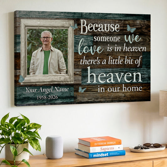 A personalized wooden wall canvas print hanging on a white wall above a wooden console table. The canvas features a rustic wood-grain background with white and light blue text that reads 'Because someone we love is in heaven there's a little bit of heaven in our home', along with a customizable photo frame containing a portrait of a deceased loved one.